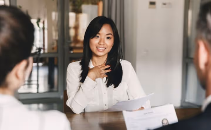 Young woman in white shirt speaking and holding papers during a job interview with two interviewers.