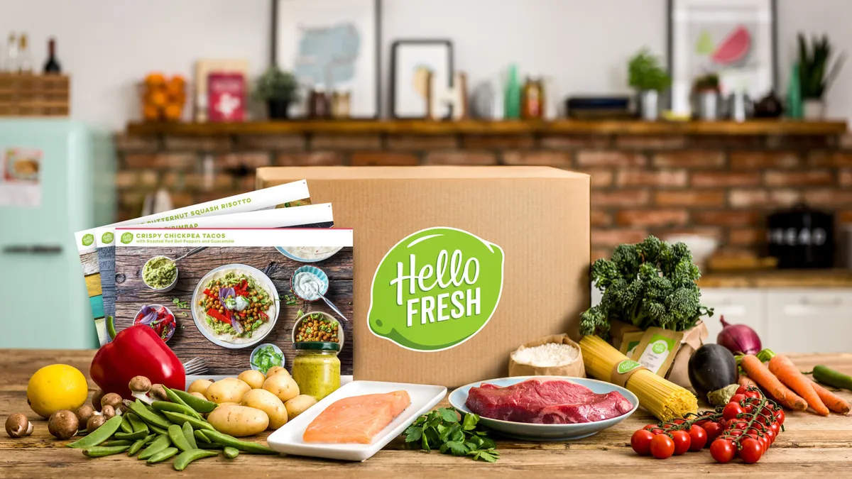 HelloFresh meal kit box surrounded by fresh ingredients including salmon, beef, cherry tomatoes, carrots, broccoli, potatoes, mushrooms, bell pepper, and recipe cards on a wooden table in a kitchen.