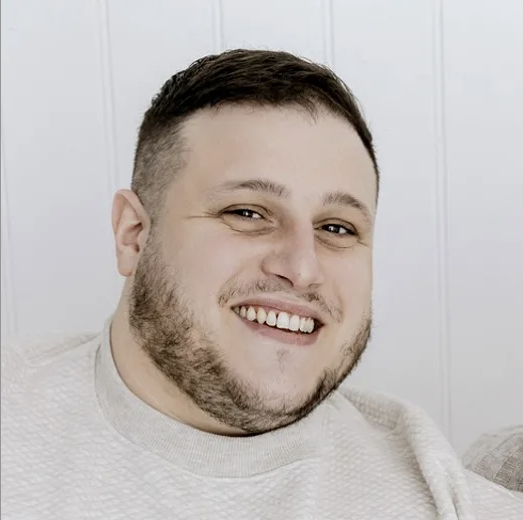Smiling man with short dark hair and beard wearing a light-colored sweater against a white background.