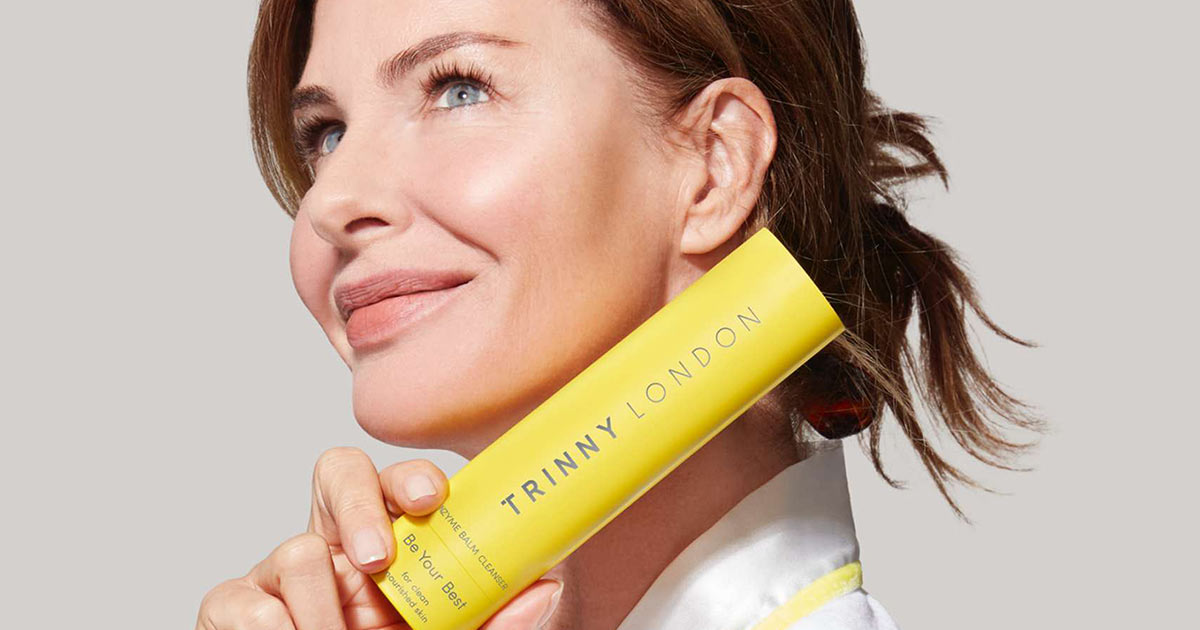 Smiling woman holding a yellow tube of Trinny London enzyme balm cleanser near her face.